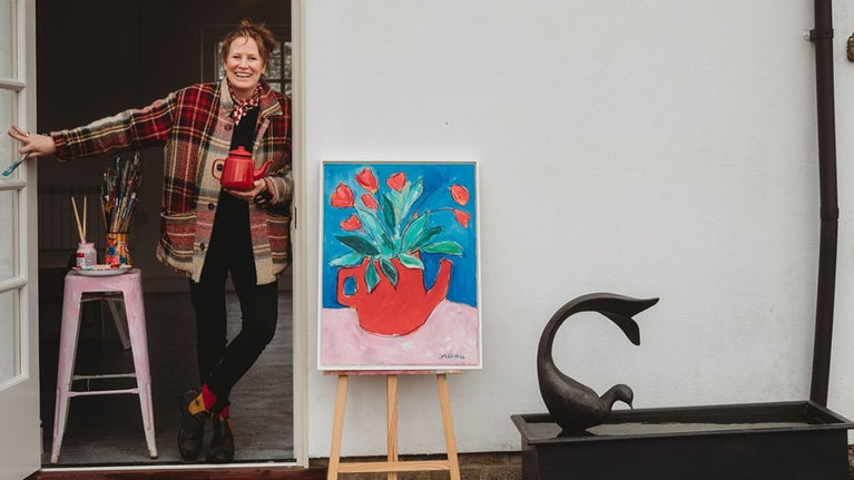 Artist Georgie Weedon posing in the doorway of the Racquets Court in Avebury Manor Garden, next to a colourful painting of a red jug containing bright red flowers against a blue backgrounfd.